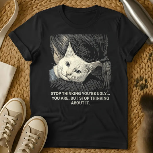 Black - A t-shirt design featuring an engraved-style illustration of a light beige cat's head with large, wide eyes and a gray collar, surrounded by swirling lines. Below the cat, the sarcastic quote reads: "STOP THINKING YOU'RE UGLY... YOU ARE, BUT STOP THINKING ABOUT IT."