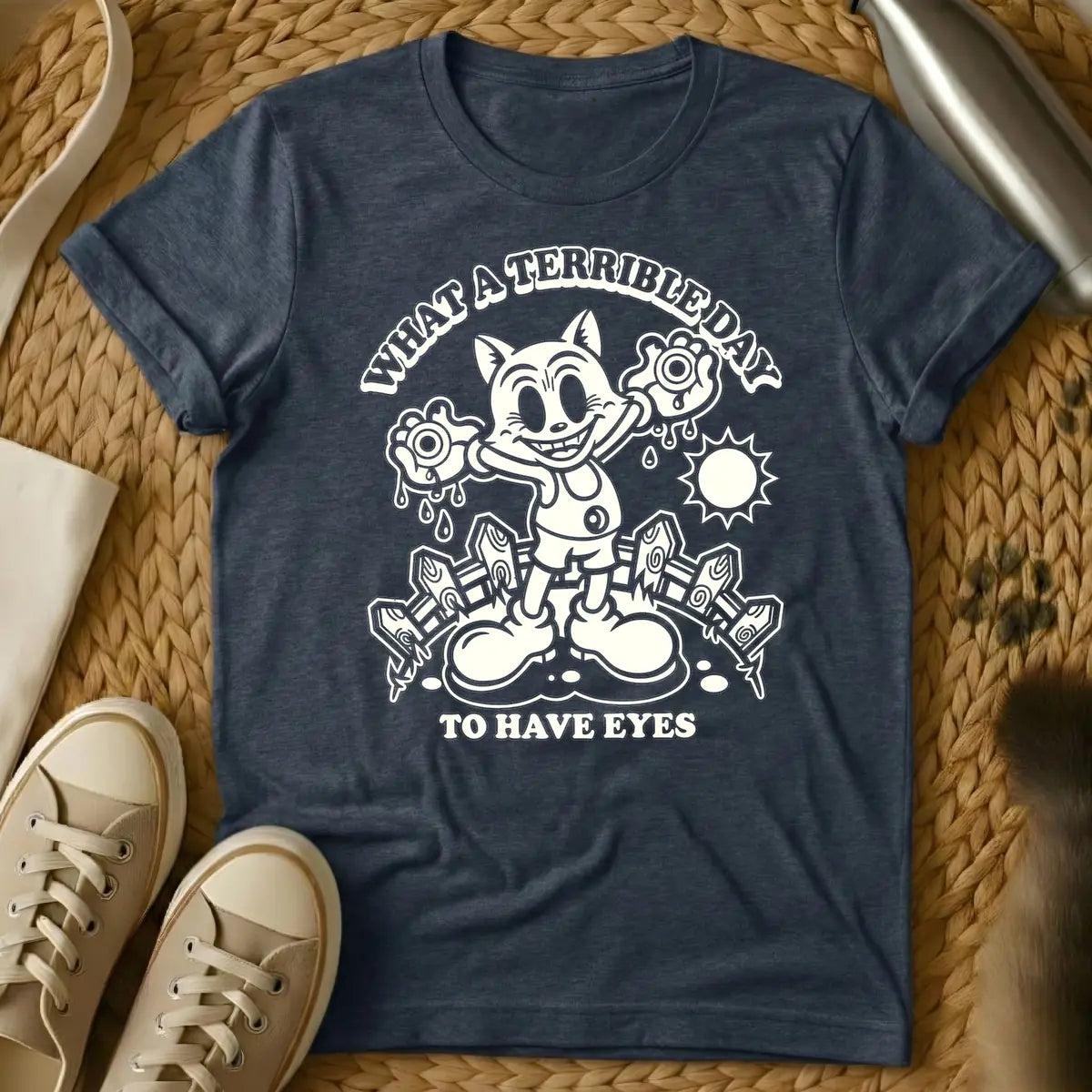 Heather Navy - Cream retro cartoon character, resembling a grinning cat with large eyes, holding two dripping eyeballs in its hands. Behind it are a picket fence and a bright sun. Arched text above reads "WHAT A TERRIBLE DAY" and straight text below reads "TO HAVE EYES".